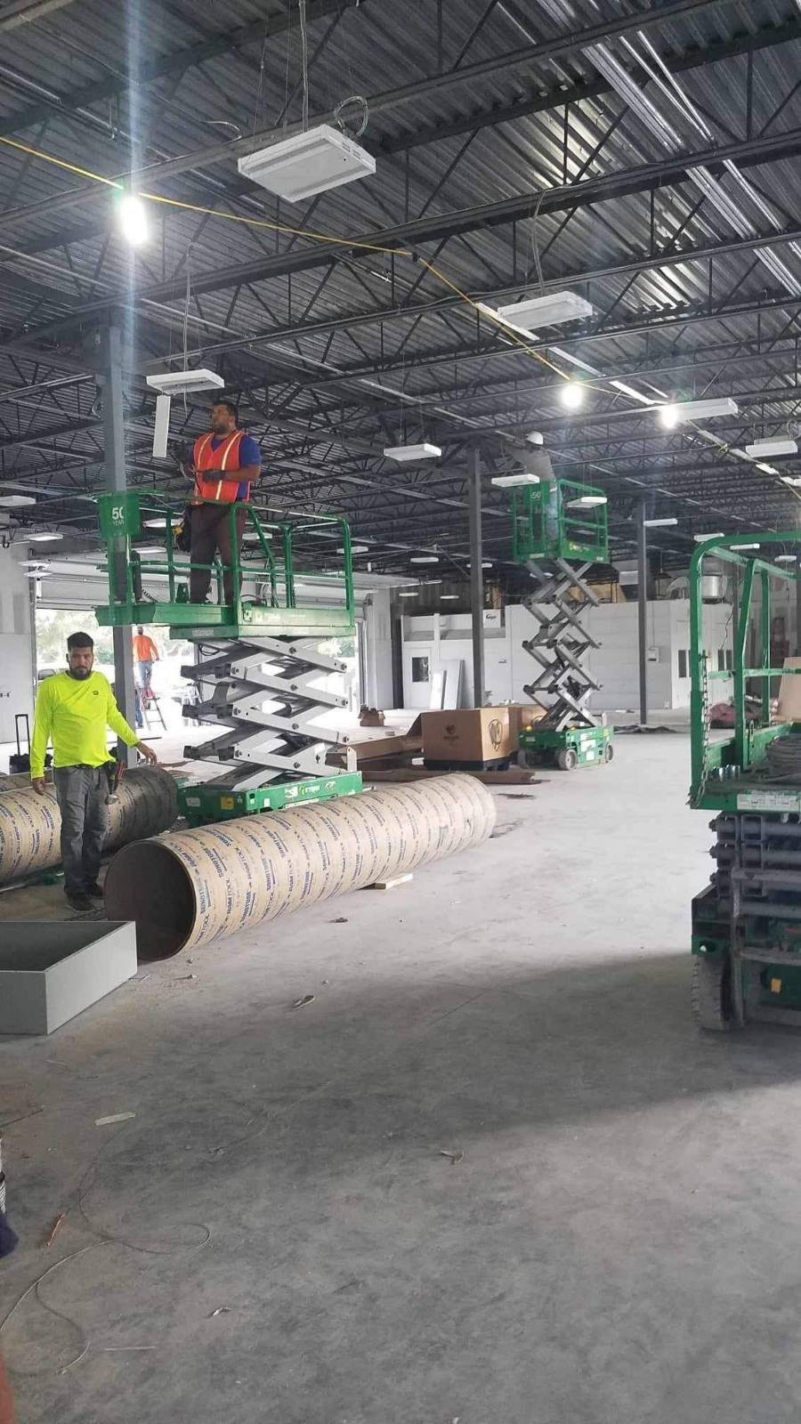 Workers in safety gear operating green scissor lifts in industrial warehouse with metal pipes.