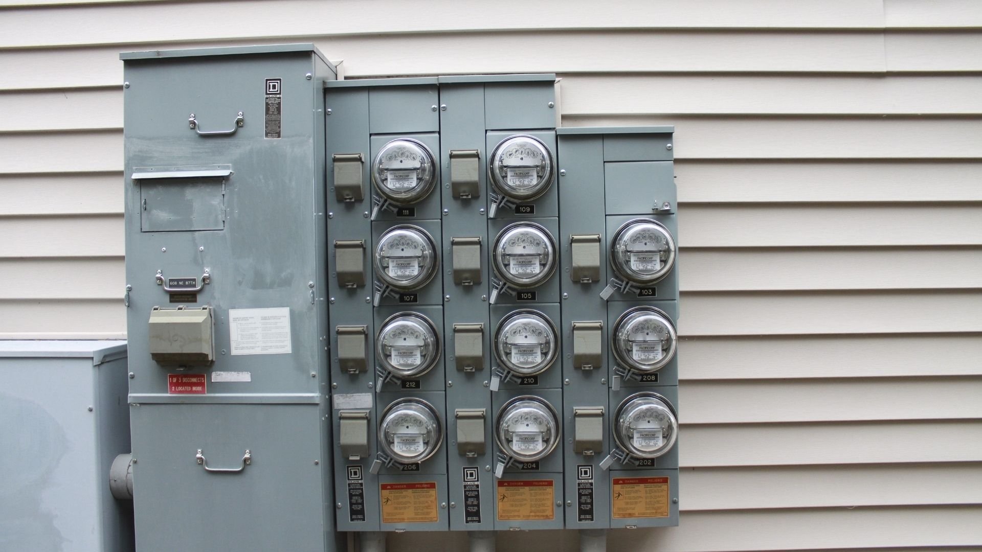 Gray electrical meter box with multiple round meters mounted on white siding wall