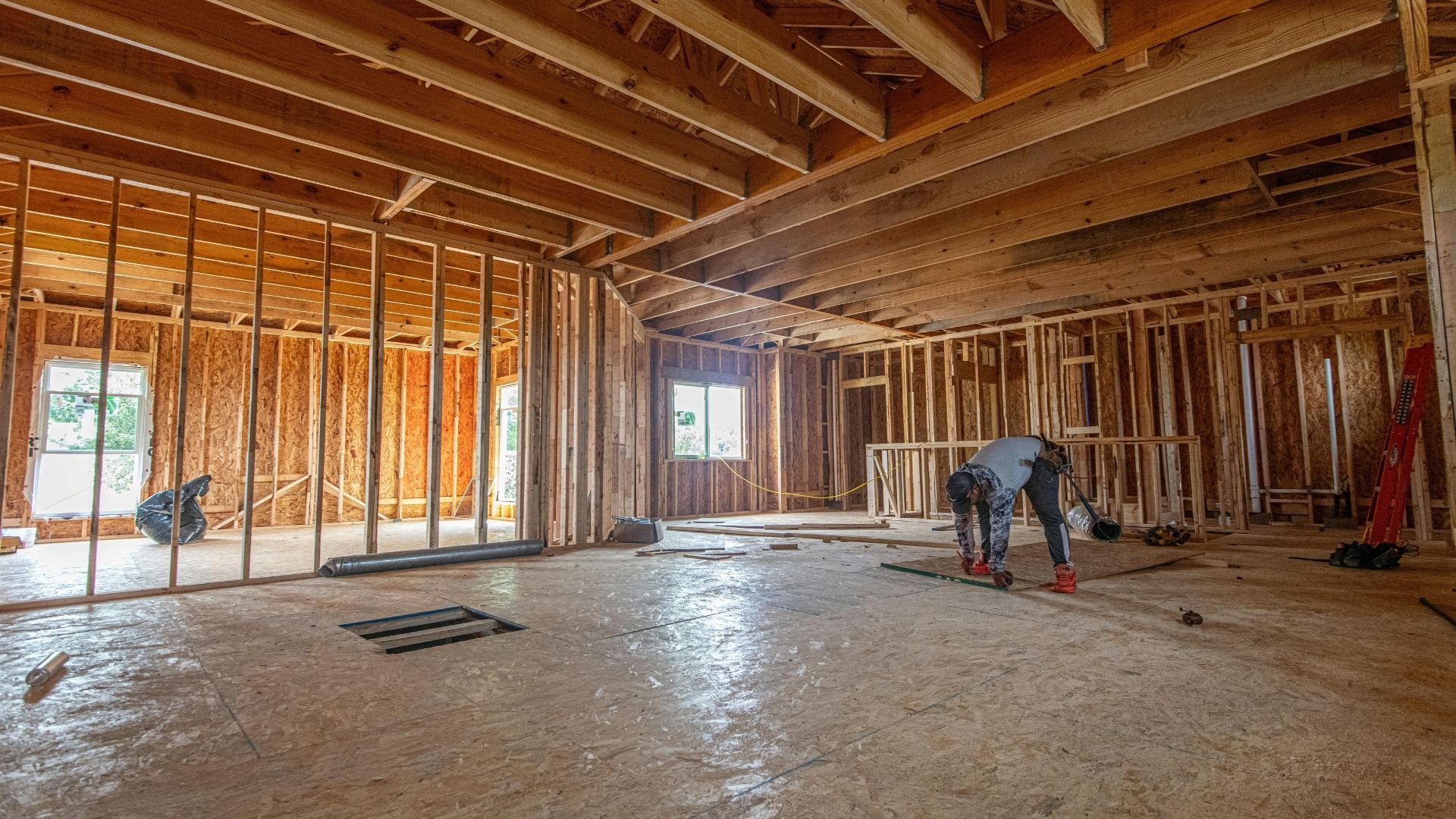 Construction worker bending in an unfinished home interior with exposed wooden framing and beams.