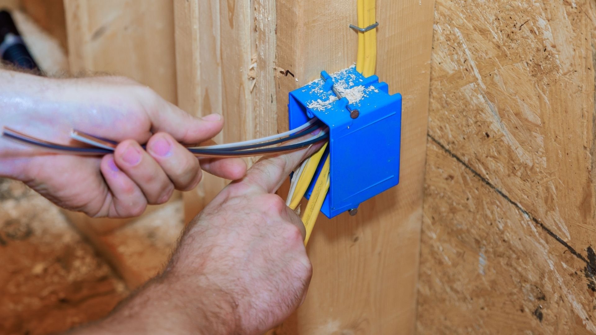 Electrician's hand inserting wires into blue electrical junction box mounted on wooden wall.