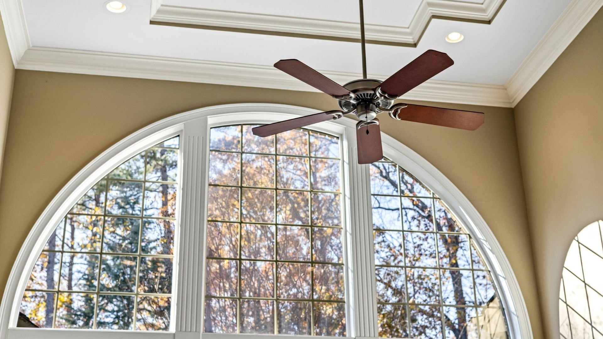 Ceiling fan with dark wood blades in front of large arched window overlooking autumn trees