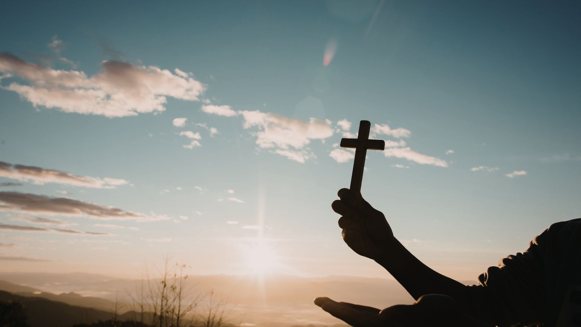 Silhouetted hand holding Christian cross against scenic sunset sky with clouds.