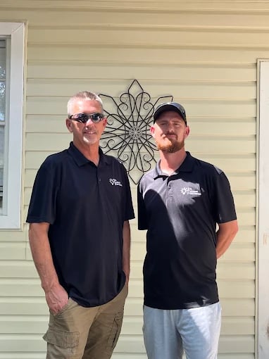 Two men in black polo shirts standing together in front of a beige house with decorative metal wall art.